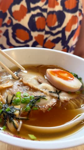 Chopsticks lift pork slice from ramen bowl, bright restaurant lighting, close-up, shallow depth