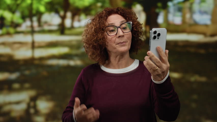 Woman in park video calling on smartphone surrounded by nature enjoying outdoor conversation in a sunny setting.