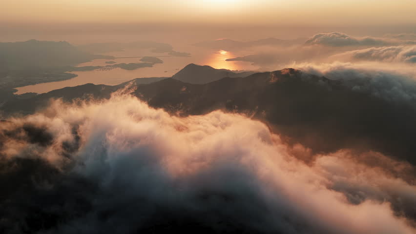 Hong Kong mountain landscape at sunrise, golden mist and clouds drifting above peaks with glowing coastline and calm sea horizon, tranquil aerial nature view ideal for travel and tourism
