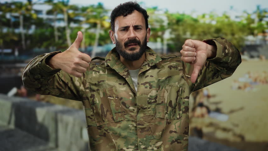 Military man with beard in camouflage uniform gestures thumbs up and down while standing in a green park with trees in the background, displaying mixed emotions outdoors.