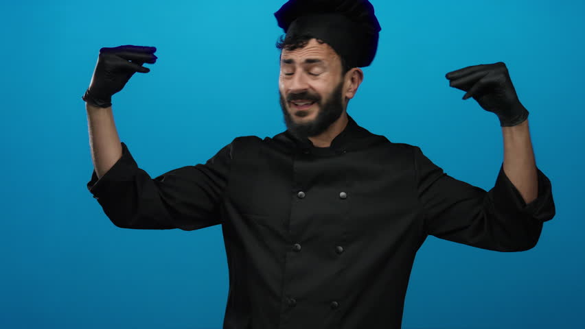 Hispanic man with beard in black chef uniform and gloves making expressive gestures against blue backdrop.