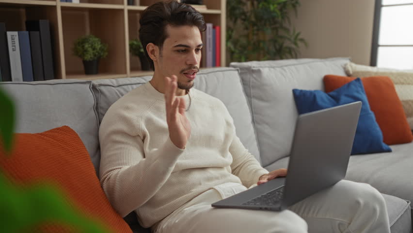 Young man sitting indoors using laptop for video call surrounded by cozy decor in modern living room setting showcasing casual relaxation and communication.