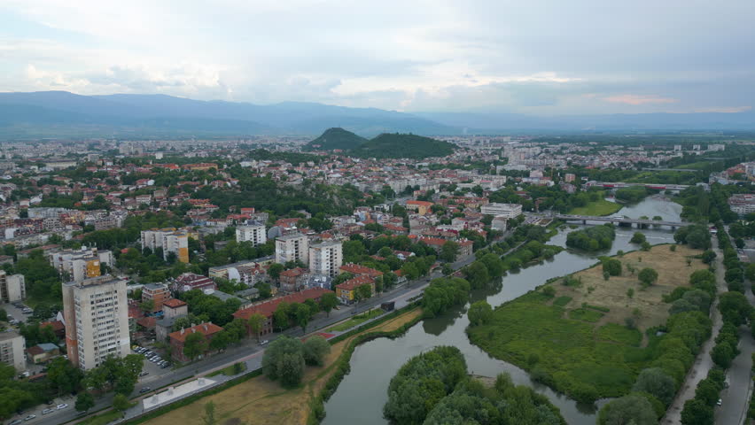 Wide aerial view of Plovdiv, Bulgaria, with the Maritsa River and bridges in the foreground, and distant hills under a cloudy spring sky at sunset.
