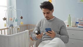 Young man in gray sweater sits in a baby's room holding a phone and medicine, surrounded by baby crib, toys, and baby care items, indicating a home and nursery setting. - Powered by Shutterstock - Get 15% off with code: PIKWIZARD15