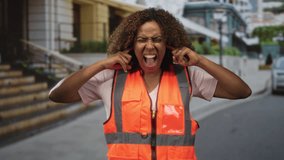 Woman in orange high visibility vest plugs fingers into ears and screams on a city street near steps and parked car; frustration safety. - Powered by Shutterstock - Get 15% off with code: PIKWIZARD15