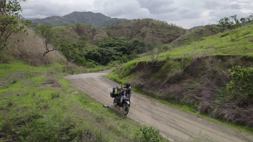 Man biker riding motorcycle, adjusting jacket, starting road trip on off-road countryside of Venezuela, South America. Aerial view captures scenic landscapes, open fields, and rural adventure.