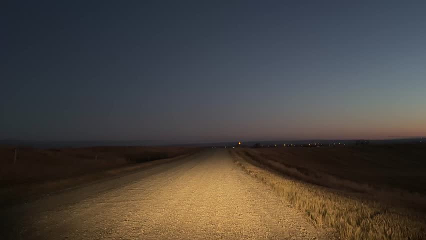 Vehicle headlights shining on a gravel country road through open farmland during nighttime in rural Alberta.
