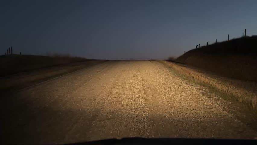 Vehicle headlights shining on a gravel country road through open farmland during nighttime in rural Alberta.