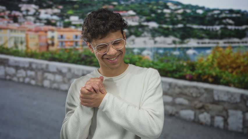 Man wearing glasses and a white sweater clasping hands on a sunlit seaside promenade with a stone wall and distant hillside village; gratitude.