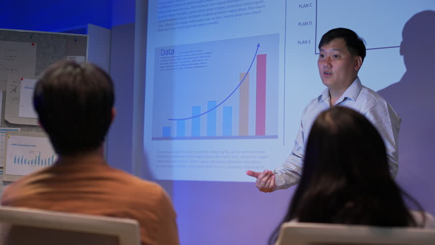 Confident Asian business professionals giving data-driven presentations to team in dark corporate office. Showing analytics charts, financial growth, and global strategy under projector light.