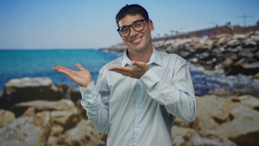 Man wearing glasses in white shirt smiles and points open palms at rocky shore and blue sea in studio; optimism.