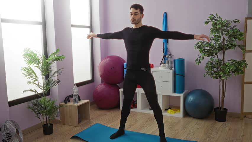 Young man stretching in gymnasium with yoga mat, plants, exercise balls, and equipment, showcasing fitness activity in a bright indoor sports center setting.