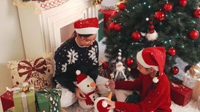 Two kids in Santa hats sit by a decorated Christmas tree, exchanging gifts and playing with snowman toys beside wrapped presents and holiday decor near a fireplace. 4k video - Powered by Shutterstock - Get 15% off with code: PIKWIZARD15