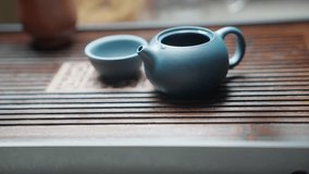 Slow motion side view: hot water is poured into a blue clay teapot on a bamboo tea tray, creating splashes and rising liquid, with a blurred blue clay teacup in the background. - Powered by Shutterstock - Get 15% off with code: PIKWIZARD15
