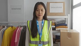Young chinese woman in reflective vest volunteering in an indoor charity center surrounded by donated clothes for community service. - Powered by Shutterstock - Get 15% off with code: PIKWIZARD15
