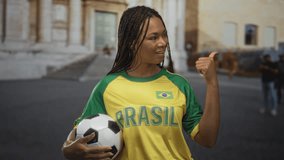 Woman holding soccer ball points thumb toward street in old town by historic building wearing brasil jersey and braids; pride. - Powered by Shutterstock - Get 15% off with code: PIKWIZARD15
