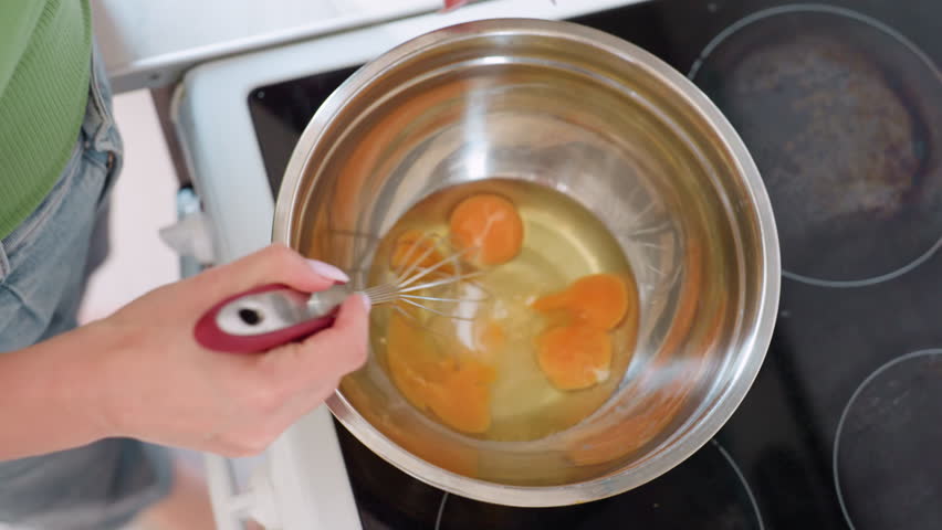 Top down view of chef whisking egg mixture in stainless steel bowl, blending yolk and white together during baking preparation process, handheld kitchen utensil mixing technique
