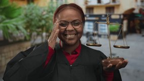 Woman judge wearing robe holding balance scales with one hand and smiling against street backdrop; justice joy. - Powered by Shutterstock - Get 15% off with code: PIKWIZARD15