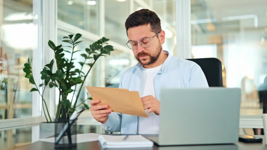Worried businessman in light blue shirt, glasses, reads document at office desk, revealing stress and disappointment. Modern office setting. Male professional experiences financial problem.