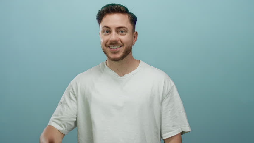 Young man smiling with a thumbs up gesture against a vibrant blue background, exuding positivity and confidence in an isolated studio setting.