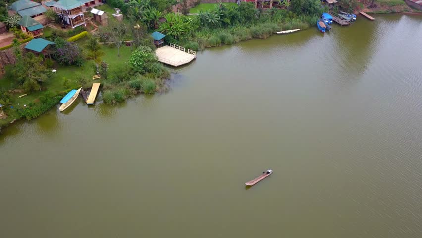 Drone view of a wooden dugout canoe on Lake Bunyonyi, Uganda, showing calm waters, shoreline vegetation, and lakeside community buildings.