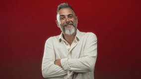 Middle aged man with arms crossed and grey beard wearing offwhite linen shirt against a studio red backdrop; confidence maturity. - Powered by Shutterstock - Get 15% off with code: PIKWIZARD15