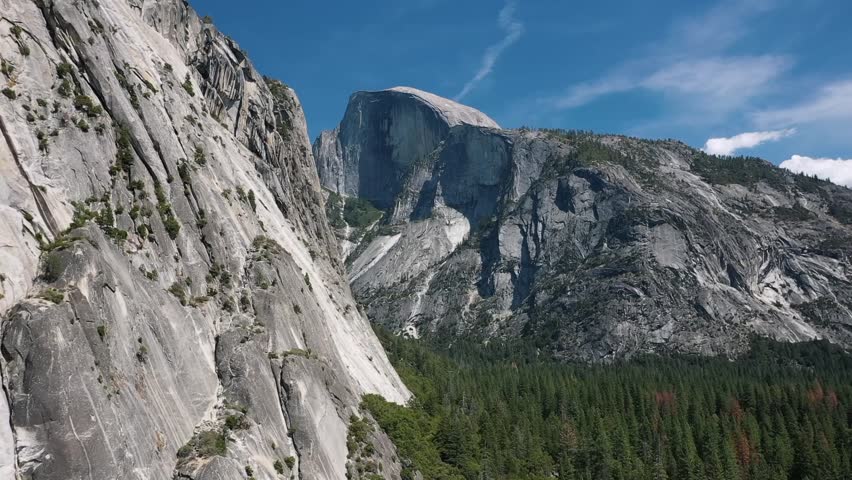 Beautiful aerial drone view showing Half Dome granite peak rising above dense forest in Yosemite National Park, California, with clear blue sky and rugged mountain terrain