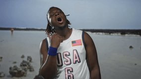 Man, young african american, mouth open and head tilted back wearing usa jersey with flag patch at beach shoreline with shallow water and rocks visible; calm reflection. - Powered by Shutterstock - Get 15% off with code: PIKWIZARD15