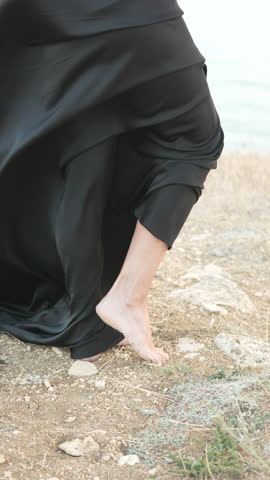 Woman, black dress, barefoot woman stands on rocky ground by the sea smiling, wind blows, vertical video