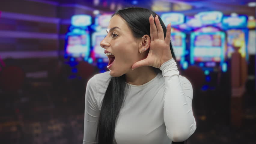 Woman smiling playfully with hand to ear in vibrant casino setting near slot machines highlighting the lively atmosphere indoors with bright lights.
