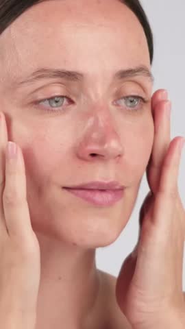 Vertical close-up of a woman gently massaging her face skin while applying face serum or facial oil. Hydrating skincare treatment and self-care concept with copy space.