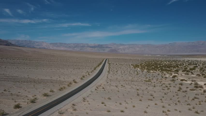 Aerial drone view showing a long straight road stretching across a dry desert landscape surrounded by sparse vegetation and distant mountains in Death Valley, California.