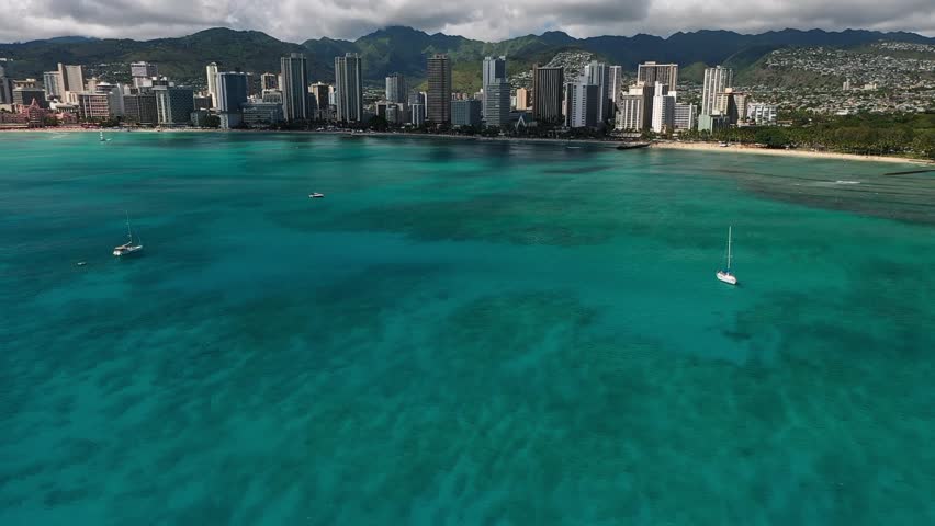 Aerial drone video showing Honolulu skyline with turquoise ocean water, sandy shoreline, and sailboats near Waikiki Beach on Oahu Island under bright sunny sky in Hawaii