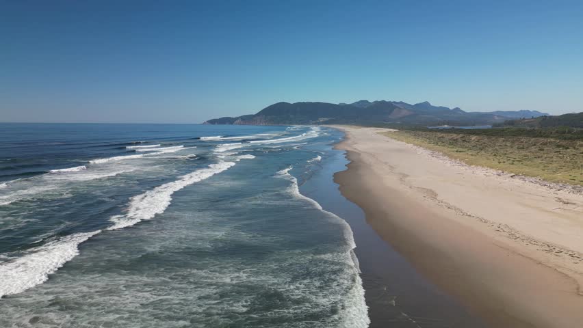 A long sandy coastline with gentle waves and mountains in the distance at Waikato, New Zealand