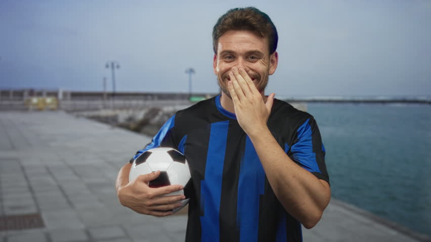 Man holding soccer ball with hand over mouth on street promenade by the sea wearing blue jersey, smiling; joy team spirit.