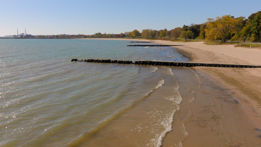 Drone aerial flying low over a concrete pier and gentle Lake Michigan waves beside the beach and shoreline road lined with colorful autumn trees in Sheboygan, Wisconsin.