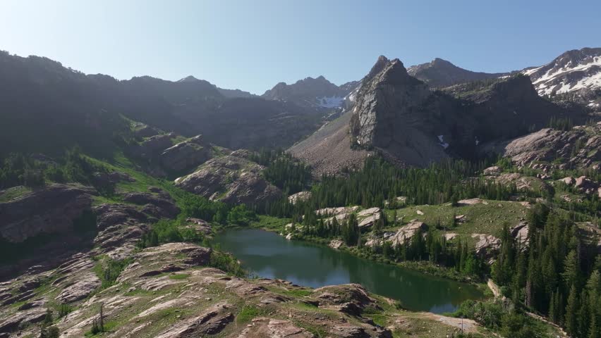 Aerial drone trucking right shot of Lake Blanche in Big Cottonwood Canyon near Salt Lake City, Utah, on a bright green summer morning with rocky peaks, lush foliage, and Sundial Peak towering above