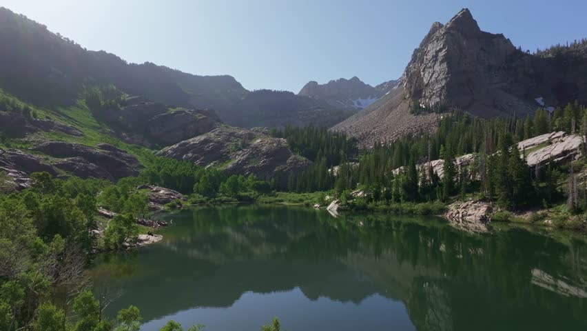 Aerial drone dolly out shot of Lake Blanche in Big Cottonwood Canyon near Salt Lake City, Utah, on a bright green summer morning with rocky peaks, lush foliage, and Sundial Peak towering above