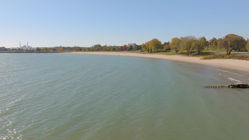 Drone aerial over Lake Michigan in Sheboygan, Wisconsin on a bright autumn day, showing beach and colorful fall trees along the shoreline under a clear blue sky.