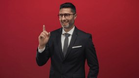 Hispanic man in suit and tie pointing both index fingers upward in red studio, wearing glasses and pocket square; confidence. - Powered by Shutterstock - Get 15% off with code: PIKWIZARD15
