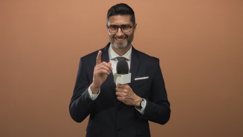 Man reporter holding microphone and pointing finger while smiling, wearing suit and tie in a peach studio; confidence professionalism.