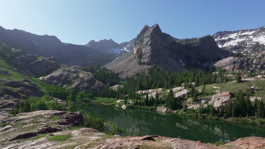 Aerial drone trucking left shot of Lake Blanche in Big Cottonwood Canyon near Salt Lake City, Utah, on a bright green summer morning with rocky peaks, lush foliage, and Sundial Peak towering above