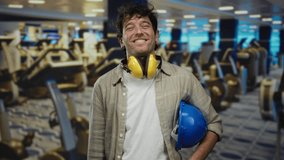 Hispanic man wearing headphones and a hardhat smiling confidently in a modern gym, surrounded by exercise equipment, highlighting indoor workplace safety. - Powered by Shutterstock - Get 15% off with code: PIKWIZARD15
