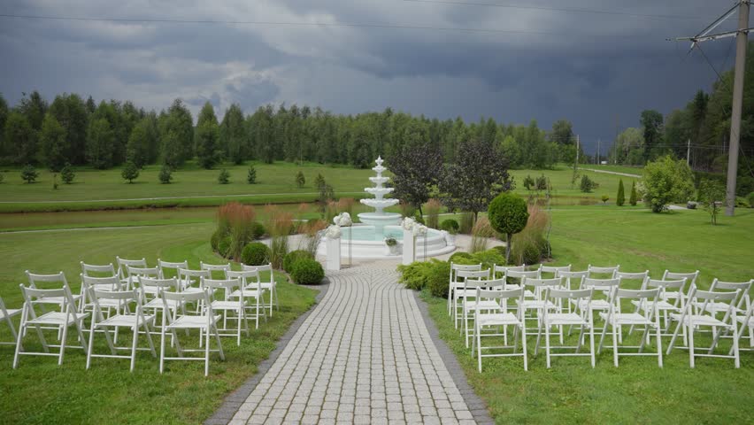 Elegant outdoor wedding ceremony setup with white chairs and decorative fountain surrounded by lush greenery under a dramatic overcast sky in a scenic countryside venue.