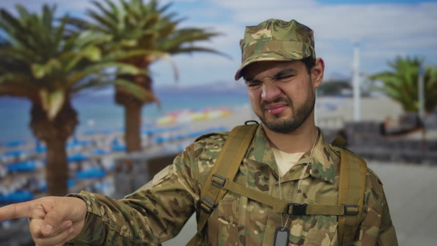 Man pinches nose on street promenade in camouflage army uniform with backpack straps and dog tags visible; disgust.