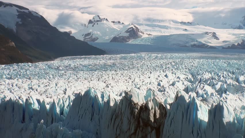 Observe a majestic glacier cascading down from the snowy mountains of Patagonia, Argentina. The view captures the immense scale and icy beauty on a cloudy day.