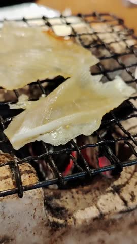 A close-up shot of thin, translucent slices of dried fish fillet (possibly stingray or skate) being grilled over hot charcoal on a black wire rack, showing the cooking process.