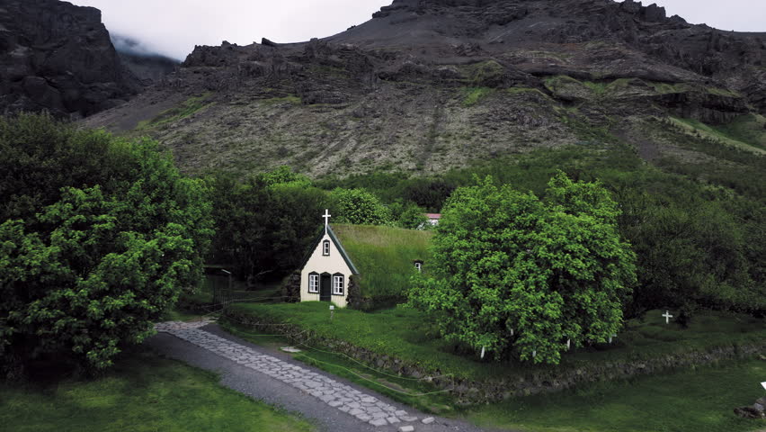 Historic turf church Hof Iceland. Aerial footage showing old wooden church with grass roof amid green landscape and volcanic hills.