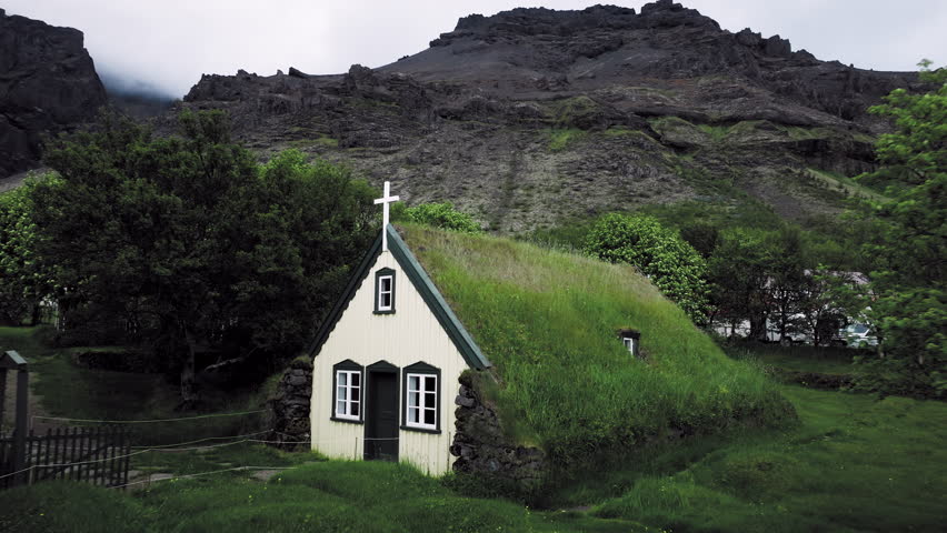 Drone shot of Hof turf church with mountain backdrop Iceland. The small wooden church sits at the foot of rocky cliffs among dense green vegetation.