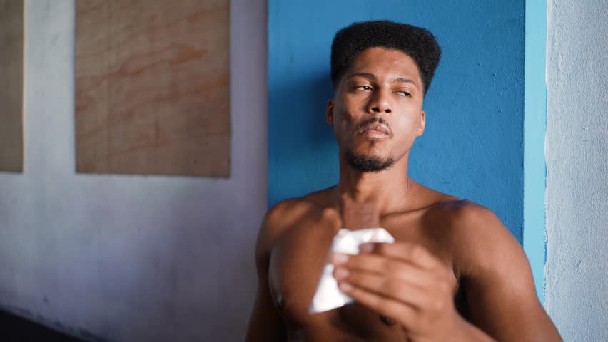 Young Black man with athletic build eating protein bar against blue wall during workout recovery period in fitness facility emphasizing post-exercise nutrition, wellness routines for muscle recovery
